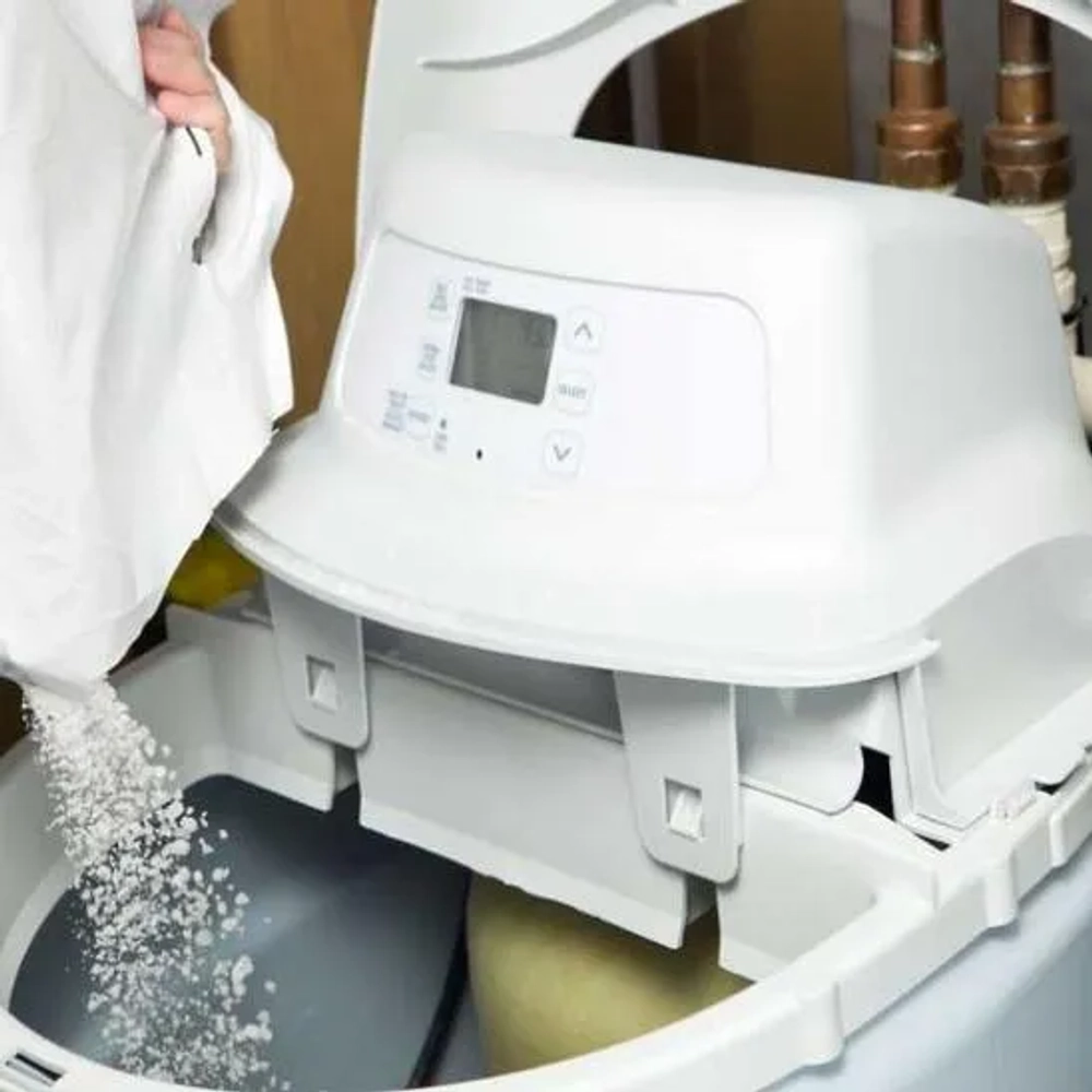 Close-up of a person refilling the brine tank of a digital water softener unit with specialized white pellet salt.