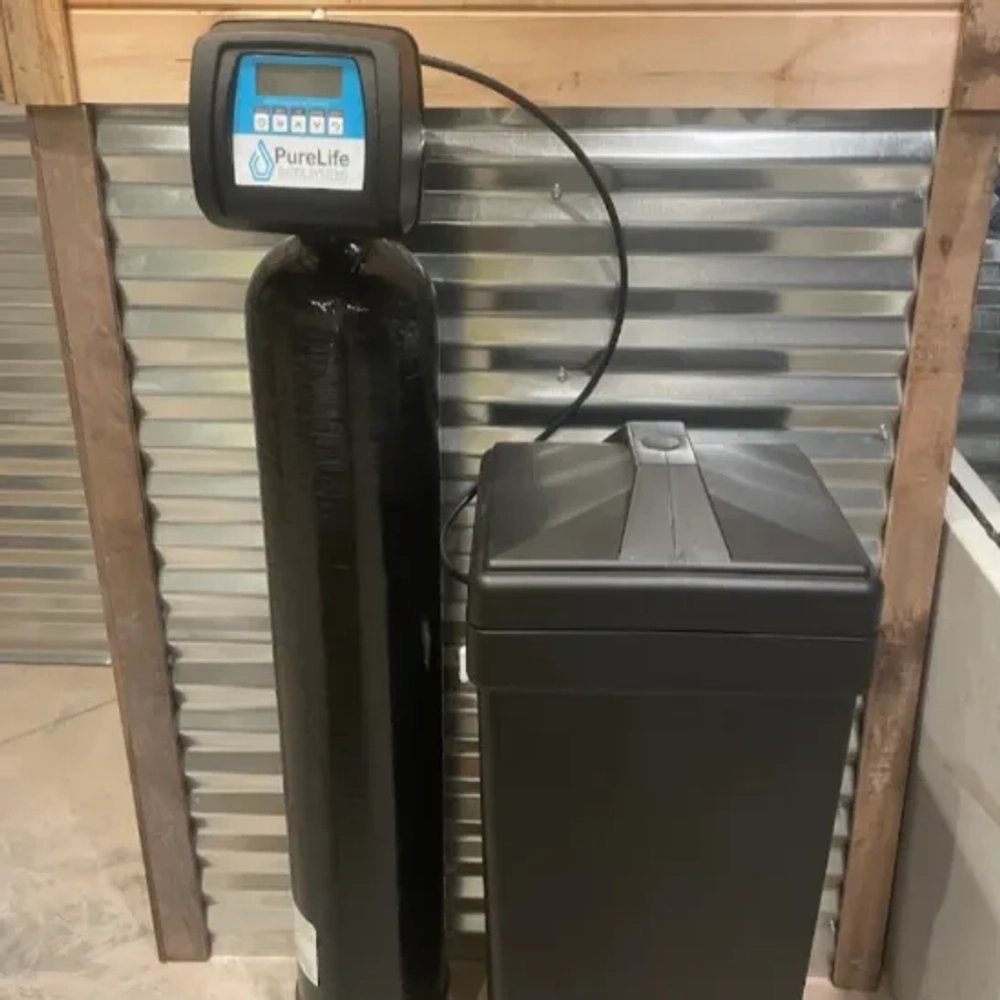 black water softener system (tank and brine tank) against a galvanized corrugated metal wall.